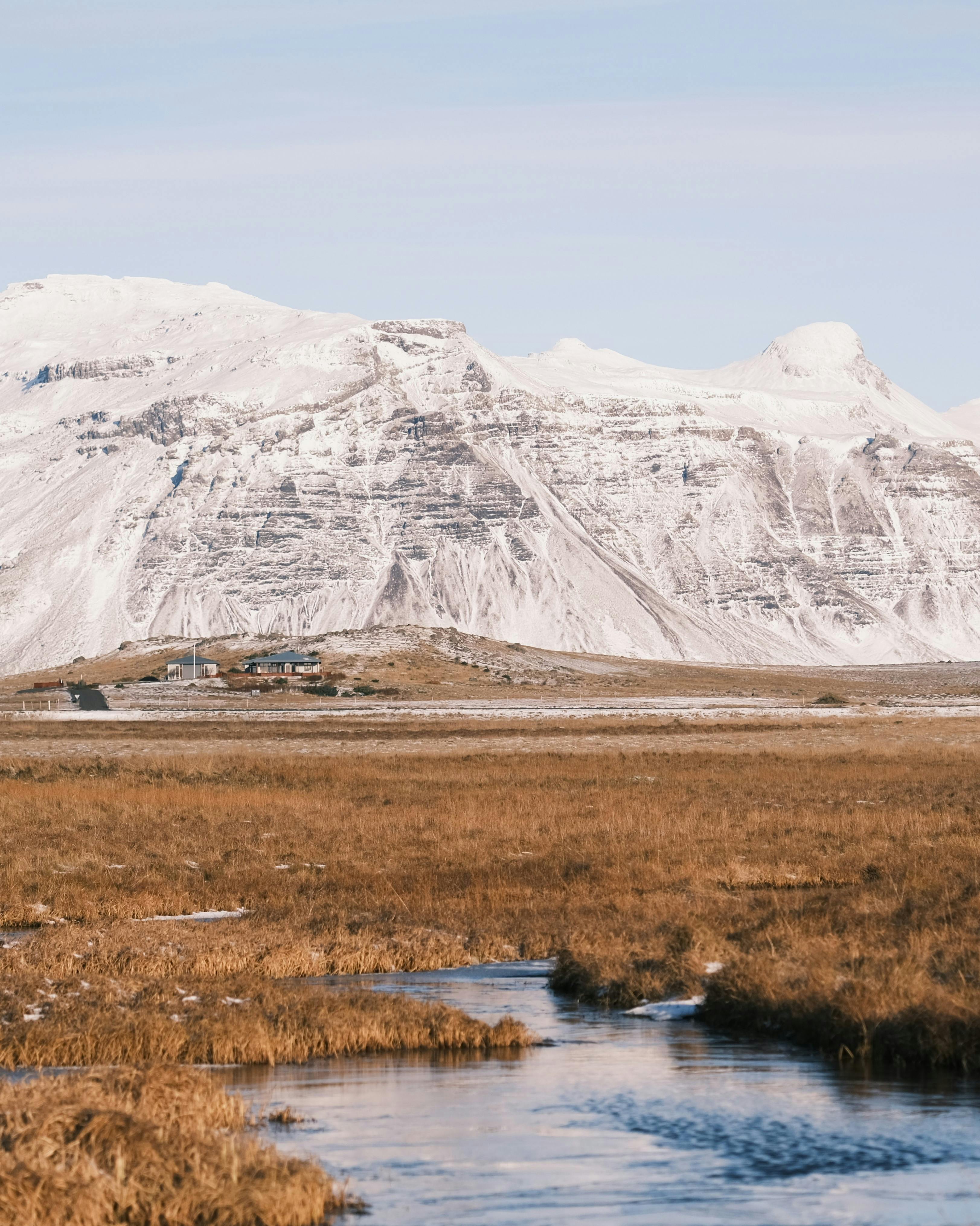 A grassy plain with a large snowy mountain behind, a small grouping of buildings in the mid ground and a stream in the foreground